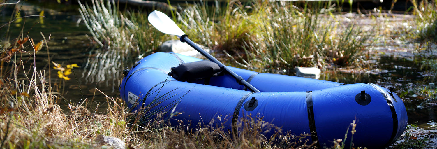 A Swellfish Jasper Packraft with a paddle is sitting on a marshy shoreline. 